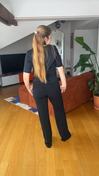 Kvinne med langt hår i et svart antrekk står i en stue med tregulv, rustfarget sofa, TV og potteplante.