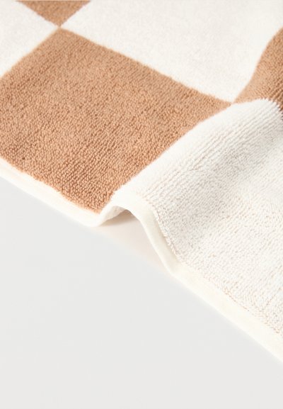 Gros plan d'une serviette beige et blanche à motif damier avec un tissu doux et texturé, partiellement pliée sur une surface blanche.