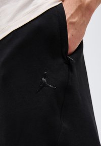 Χέρι στην τσέπη μαύρου παντελονιού με ανάγλυφο λογότυπο Jumpman κοντά στη ραφή κοντά σε λευκό πουκάμισο.