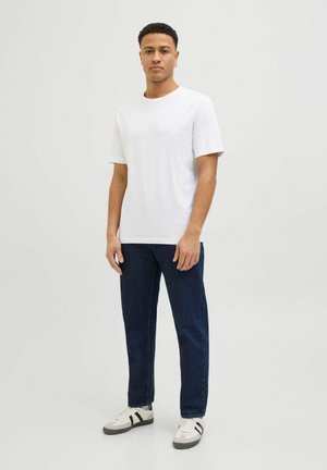 Uomo in piedi che indossa una semplice maglietta bianca a maniche corte, jeans blu scuro e sneakers bianche con strisce nere su uno sfondo chiaro uniforme.