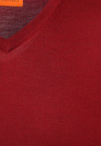 Roter Strickpullover mit V-Ausschnitt und feinem Rippstrick. Sichtbare Nahtdetails am Halsausschnitt; weiche Textur, schlichtes Design, keine Grafiken oder Muster.