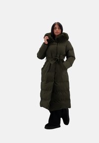 Cappotto imbottito lungo verde oliva con cappuccio foderato in pelliccia sintetica, vita con lacci e texture trapuntata. Il modello indossa pantaloni neri e calzature scure.