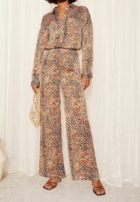 Completo in due pezzi con stampa leopardata in tessuto leggero. Camicia a maniche lunghe e pantaloni a gamba larga con motivi beige, marroni e neri. Dettagli includono polsini della camicia aderenti con bottoni e tacchi a fascia.
