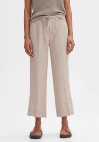 Pantalons larges en lin beige avec deux poches avant, taille à cordon et texture subtile. Associés à des sandales brunes à lanières.