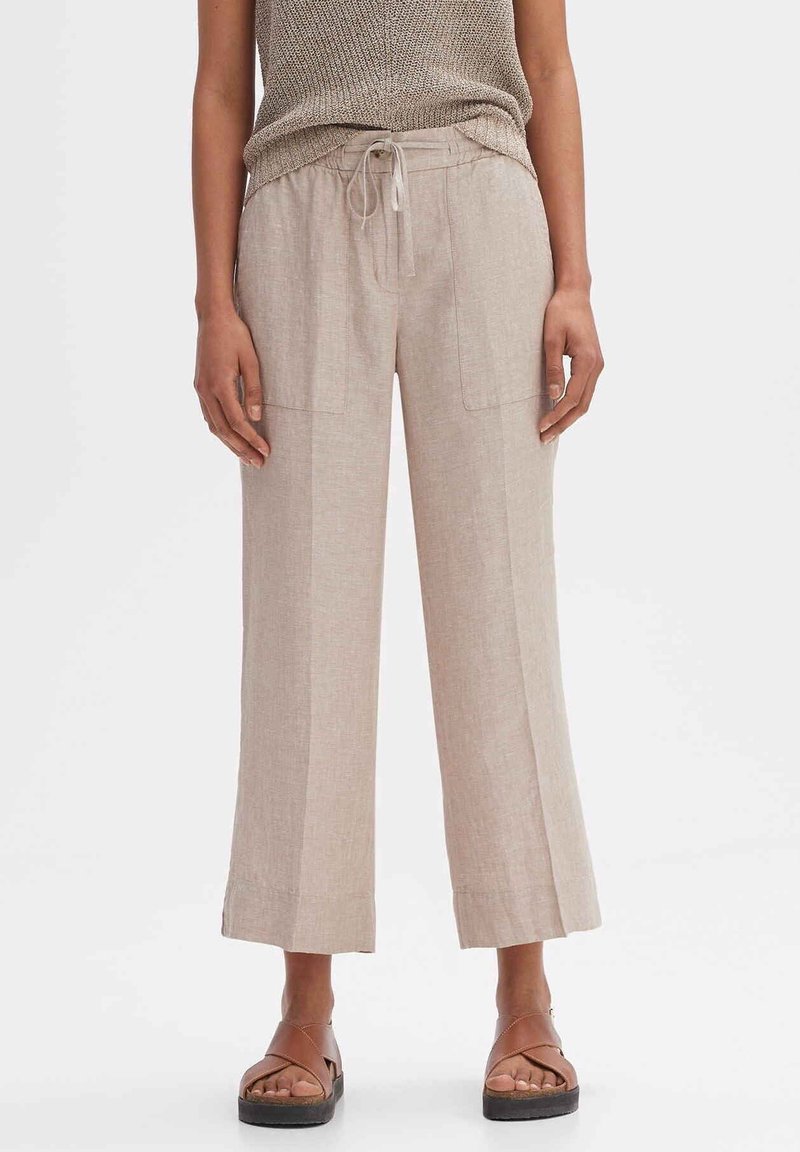 Pantalons larges en lin beige avec deux poches avant, taille à cordon et texture subtile. Associés à des sandales brunes à lanières.