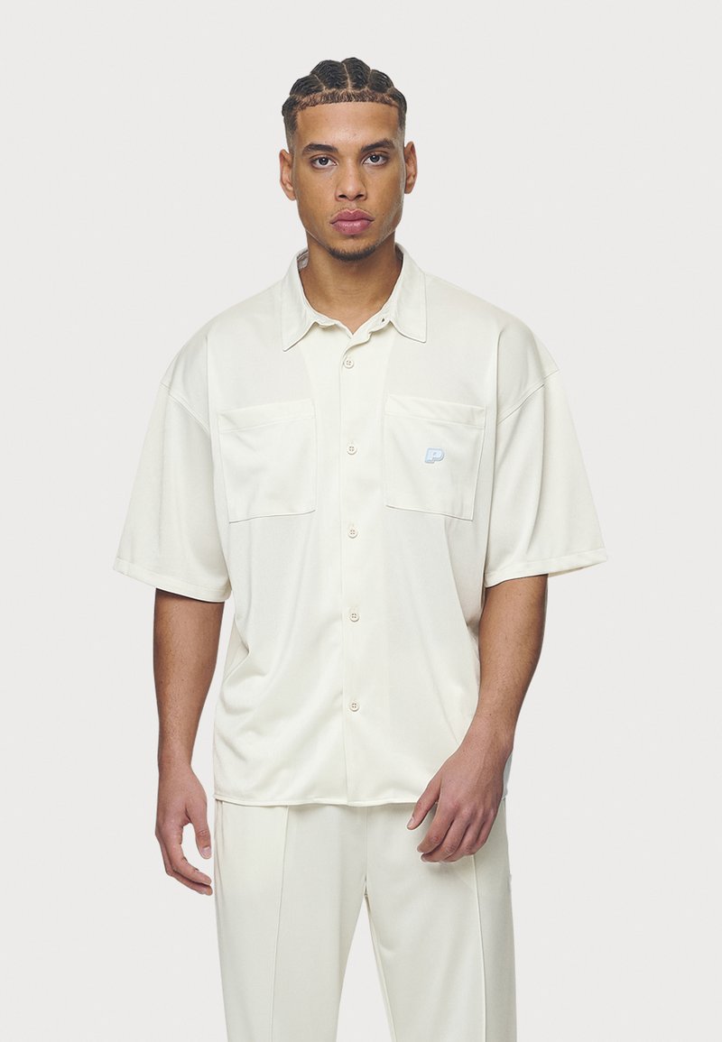 Camisa cremosa de manga corta en tejido suave con dos bolsillos frontales y un parche con el logo en el bolsillo izquierdo. Corte ajustado con un diseño relajado.