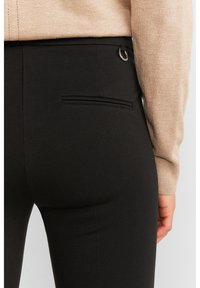 Mustat housut, joissa on sileä pinta, varustettu takataskulla ja huomaamattomilla ompeluyksityiskohdilla. Kuitu on joustavaa ja vartalonmyötäistä.