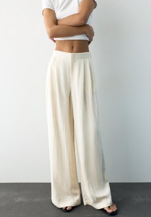 Oseba, oblečena v kremne hlače z visokim pasom in širokimi hlačnicami, črne sandale in bel kratki zgornji del s kratkimi rokavi, stoji z rokami prekrižanimi.