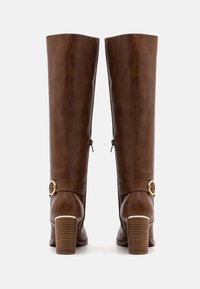 Botas marrones hasta la rodilla hechas de cuero sintético liso. Presentan un acento dorado en el tobillo y una suela de tacón apilado.