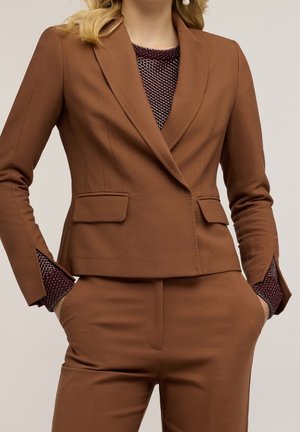 Kvinde iført brun skræddersyet jakkesæt med tætsiddende blazer og matchende bukser, hænderne i lommerne, og bordeauxmønstret top synlig ved manchetter og halsudskæring.