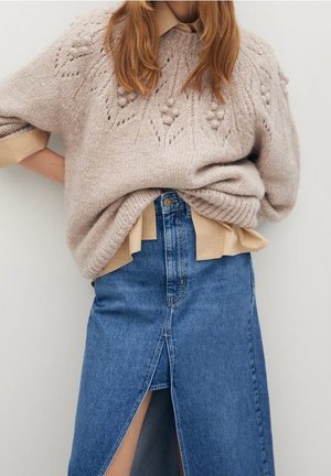 Mujer con suéter beige tejido con textura sobre una camisa color canela, combinada con una falda vaquera azul de cintura alta que presenta una abertura frontal.