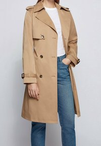 Beżowy płaszcz trencz o fasonie dwurzędowym, z dużym kołnierzem i ozdobnymi guzikami. Posiada regulowane mankiety i luźny krój. Nosi się go na białą koszulę.