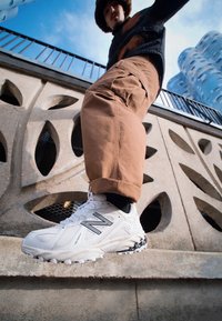 Vita sportskor i vitt med svarta detaljer, texturerat mesh överdel och grovt yttersula. Bärs tillsammans med ljusbruna cargobyxor framför en betongbakgrund.