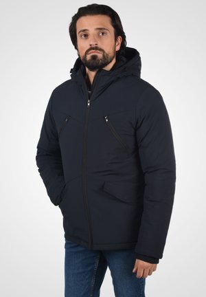 Marineblå polstret jakke med hætte, som har lynlåslomme på brystet, sidelommer og strikkede manchetter for ekstra varme og komfort.