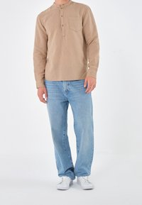 Beige langslievende henley shirt met een borstzak, gecombineerd met lichtblauwe straight-leg jeans en witte sneakers. Strak, minimaal ontwerp.