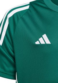 Camiseta deportiva verde con rayas blancas en los hombros y un logo bordado en blanco. Fabricada con tela suave y con un cuello redondo.