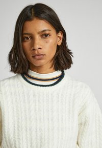 Maglione bianco a maglia con colletto rotondo, caratterizzato da strisce verde scuro, arancione e bianche. Texture morbida e vestibilità rilassata.