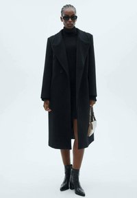 Lungo cappotto nero con ampie punte, botttoni doppi e silhouette aderente. Indossato sopra un vestito nero a collo alto e stivali alla caviglia.