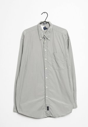 Chemise gris clair à manches longues boutonnée avec col boutonné et poche poitrine suspendue sur un cintre noir sur fond blanc.