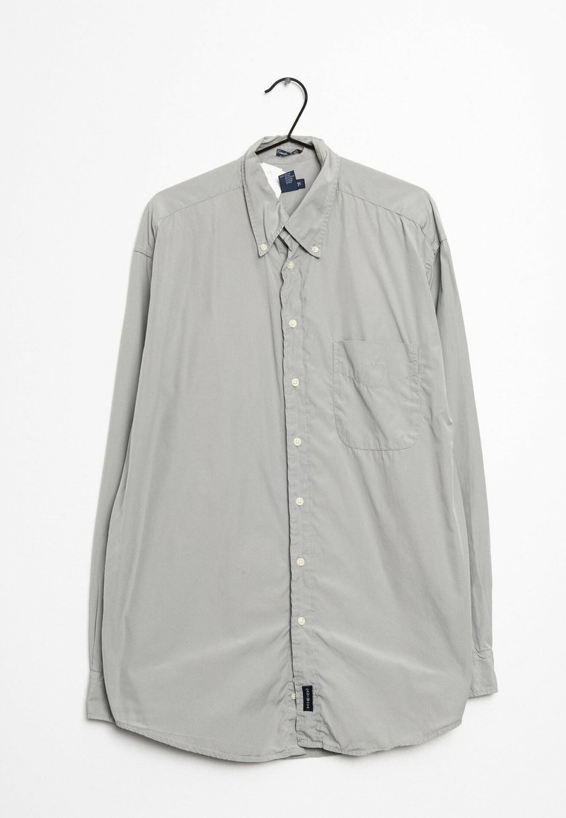 Chemise gris clair à manches longues boutonnée avec col boutonné et poche poitrine suspendue sur un cintre noir sur fond blanc.