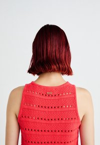 Rode gebreide tanktop met opengewerkt ontwerp. Kenmerkt een ronde halslijn en een label op de achterkant. Haar is schouderlang en glanzend.