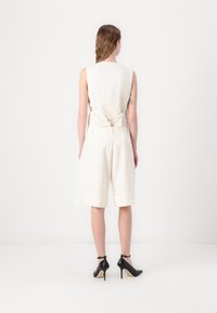 Bílý bezrukávový top s mašlí na zadní straně, v kombinaci s bílými šortkami až ke kolenům a černými vysokými podpatky.