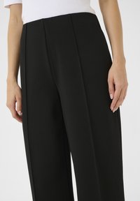 Schwarze Hosen mit glatter Textur, tiefen Falten, tailliertem Design und hoher Taille. Sichtbare Hände halten den Bund.