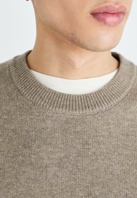 Pull en tricot marron clair avec un col ras du cou côtelé, superposé à une chemise blanche. La texture semble douce et confortable.