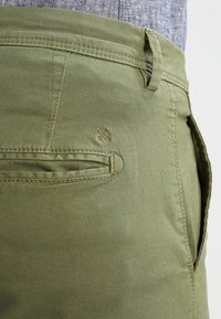 Närbild av olivgröna byxor med ficka med sned öppning, bältesloop och en liten broderad symbol med korsade pilar nära fickans söm.