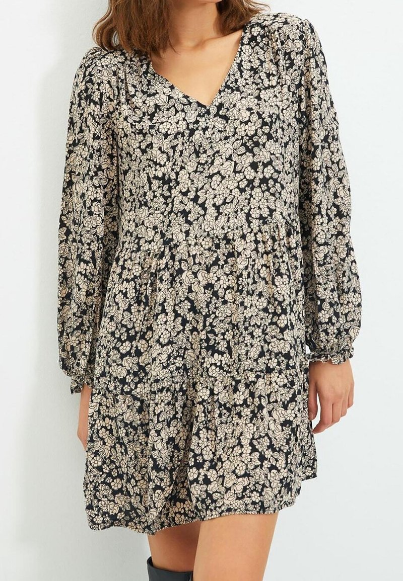 Robe noire à fleurs avec manches longues, encolure en V, taille froncée et fleurs beiges. Fabriquée en tissu léger à la texture douce.