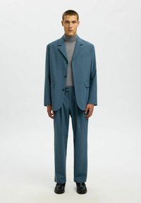 Svetlo modra oblečena kostim, ki vključuje enoredni jopič in hlače ravnega kroja, dopolnjen s sivim puloverjem s kratkim vratom in črnimi čevlji.