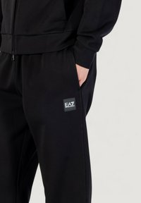 Schwarzes Zip-Up-Sweatshirt und passende Hose, hergestellt aus einem strukturierten Stoff und mit einem kleinen EA7-Logo-Patch auf der Hose.