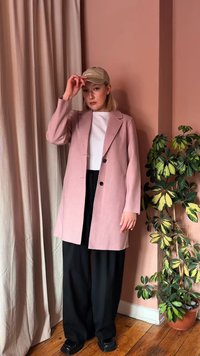 Νεαρό άτομο με μπεζ καπέλο, ανοιχτό ροζ παλτό, λευκό πουκάμισο και μαύρο παντελόνι στέκεται δίπλα σε ένα πολύχρωμο γλαστρικό φυτό με πράσινα και κίτρινα φύλλα.