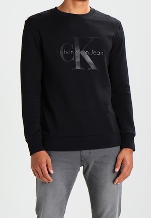 Mand iført sort Calvin Klein Jeans sweatshirt med lange ærmer og stort "CK"-logo samt grå jeans mod en ensfarvet baggrund.