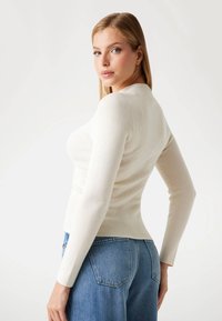 Vaalea nainen kääntyy hieman neutraalilla ilmeellä, pukeutuneena istuvaan off-white pitkähiharaiseen puseroon ja vyötäröltä korkeisiin sinisiin farkkuihin.