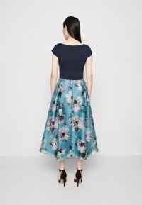 Marinblå figurnära topp med korta ärmar; ljusblå kjol med blommönster, midilängd, flödande tyg, svarta klacksandaler.