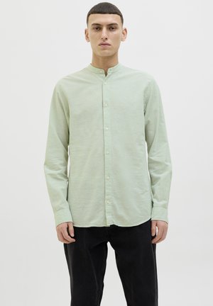 Camicia verde chiaro a maniche lunghe in un tessuto morbido, con colletto a fascia e chiusura con bottoni; indossata con pantaloni neri.