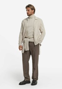 Hellbeige, kabelgestrickte Strickjacke über einem beigen Rollkragenpullover, kombiniert mit braunen gemusterten Hosen und schwarzen Schuhen. Minimalistisches Design und Textur.