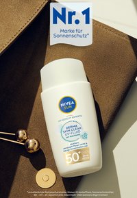 NIVEA SUN UV Fluid in einer weißen Flasche mit blauen und gelben Akzenten, ausgestattet mit einem Drehverschluss. Auf einer braunen strukturierten Oberfläche platziert.