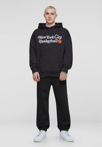 Fekete kapucnis pulóver "New York City Basketball" felirattal, kenguruzsebbe, bordázott ujjak, párosítva matching fekete melegítőnadrággal és fehér sportcipővel.