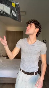 Ung mand med krøllet hår kaster en sort pakke i et soveværelse med en seng og et træskab i baggrunden.