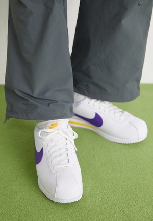 Sneakers bianche Nike con logo swoosh viola e dettagli gialli indossate con pantaloni grigio scuro su pavimento verde.