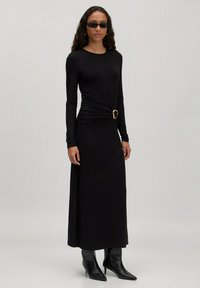 Zwarte lange mouwen jurk met een gedraaide voorkant en gouden accent. Model draagt stijlvolle zwarte zonnebril en enkelhoge laarzen. Gladde textuur.