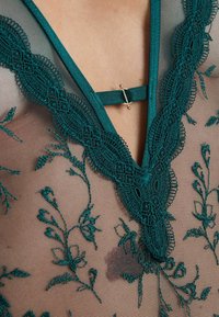 L'abito in pizzo teal con ricami floreali su rete trasparente. Presenta una scollatura a V decorativa e un dettaglio in hardware dorato.