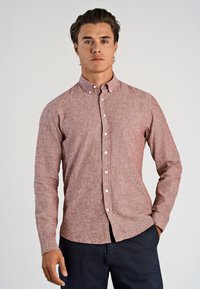 Man med brunt hår iklädd en långärmad, knäppt röd linne skjorta och mörka byxor, står med en hand i fickan mot en enkel bakgrund.