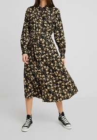 Musta kukallinen midi mekko pitkähihaisilla, napitettavalla edessä ja vyötäröllä vyöllä, jossa on kultaisia ja valkoisia kukkakuoseja. Yhdistetty mustiin lenkkareihin.