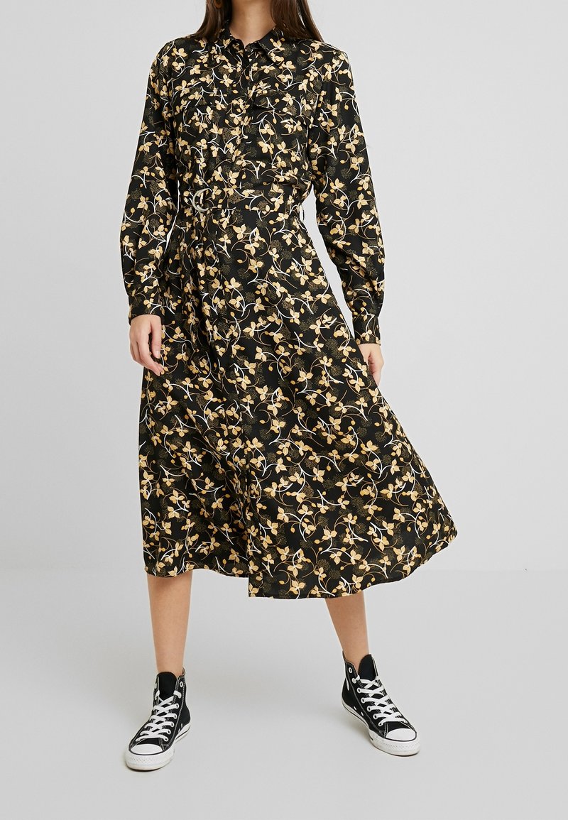 Musta kukallinen midi mekko pitkähihaisilla, napitettavalla edessä ja vyötäröllä vyöllä, jossa on kultaisia ja valkoisia kukkakuoseja. Yhdistetty mustiin lenkkareihin.