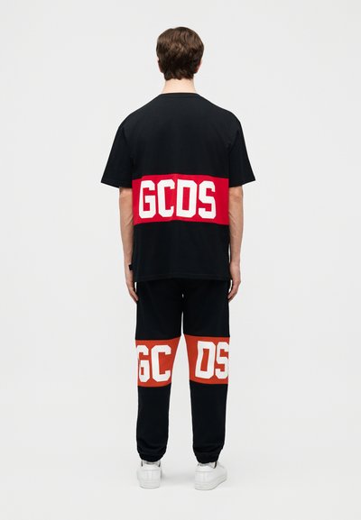 Person stående med ryggen mod kamera iført sort outfit med fed hvid "GCDS"-logo på røde bånd tværs over ryggen og de nederste ben, med hvide sneakers.