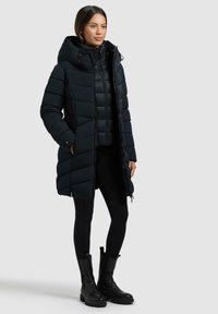 Schwarze Daunenjacke mit Kapuze, gestepptem Muster, Reißverschluss und Kordelzügen. Getragen über schwarzen Leggings und klobigen schwarzen Stiefeln.
