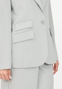 Blazer grigio chiaro con chiusura a un bottone, due grandi tasche a pattina e una trama di tessuto liscia. Vestibilità sartoriale con linee pulite.